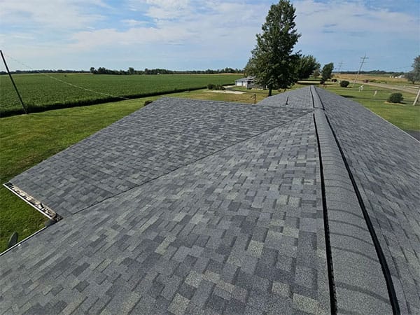 New gray architectural shingle roof with ridge cap, viewed from above with farm fields and trees beyond