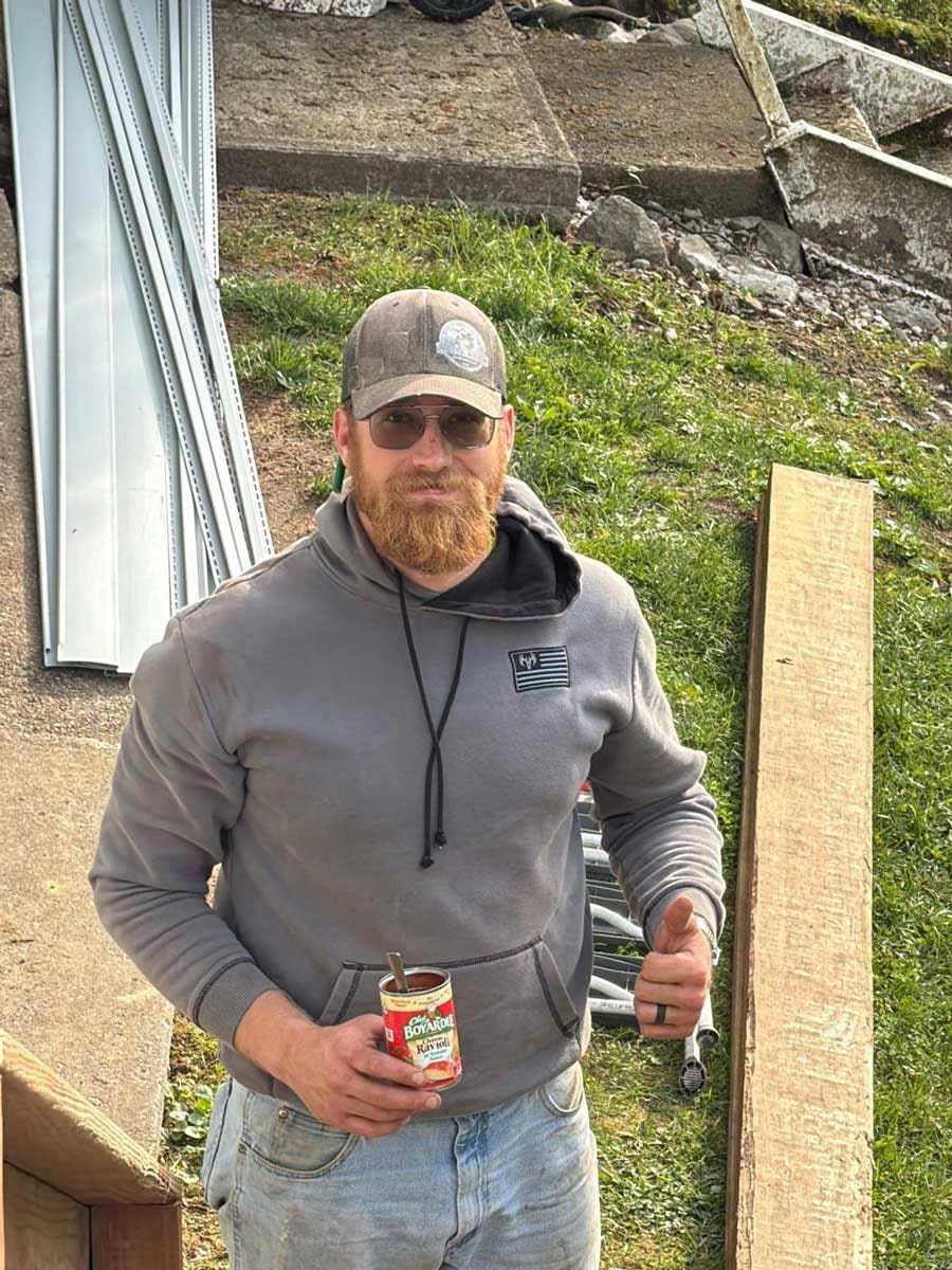 Bearded crew member in a cap and tinted glasses gives a thumbs-up on a job site while holding a can of ravioli
