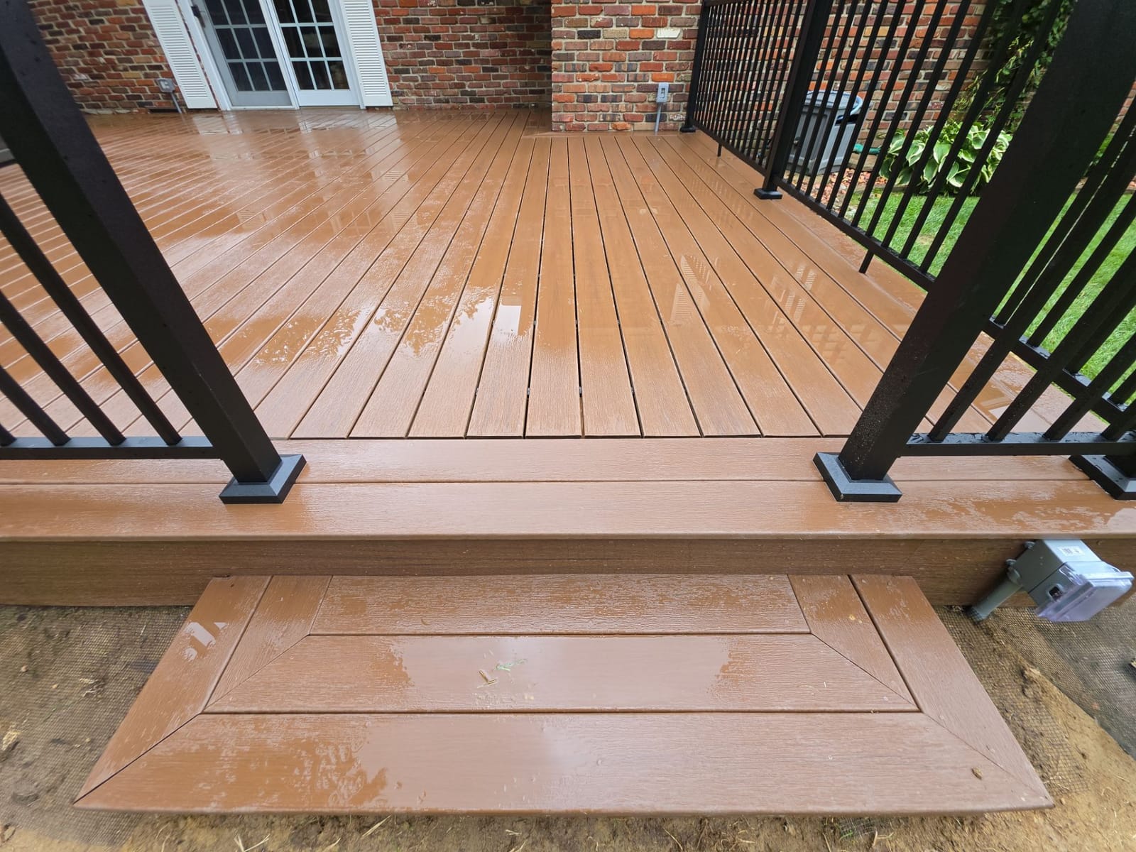 Brown composite deck with picture-frame border step and black aluminum railing posts, wet after rain, brick house behind