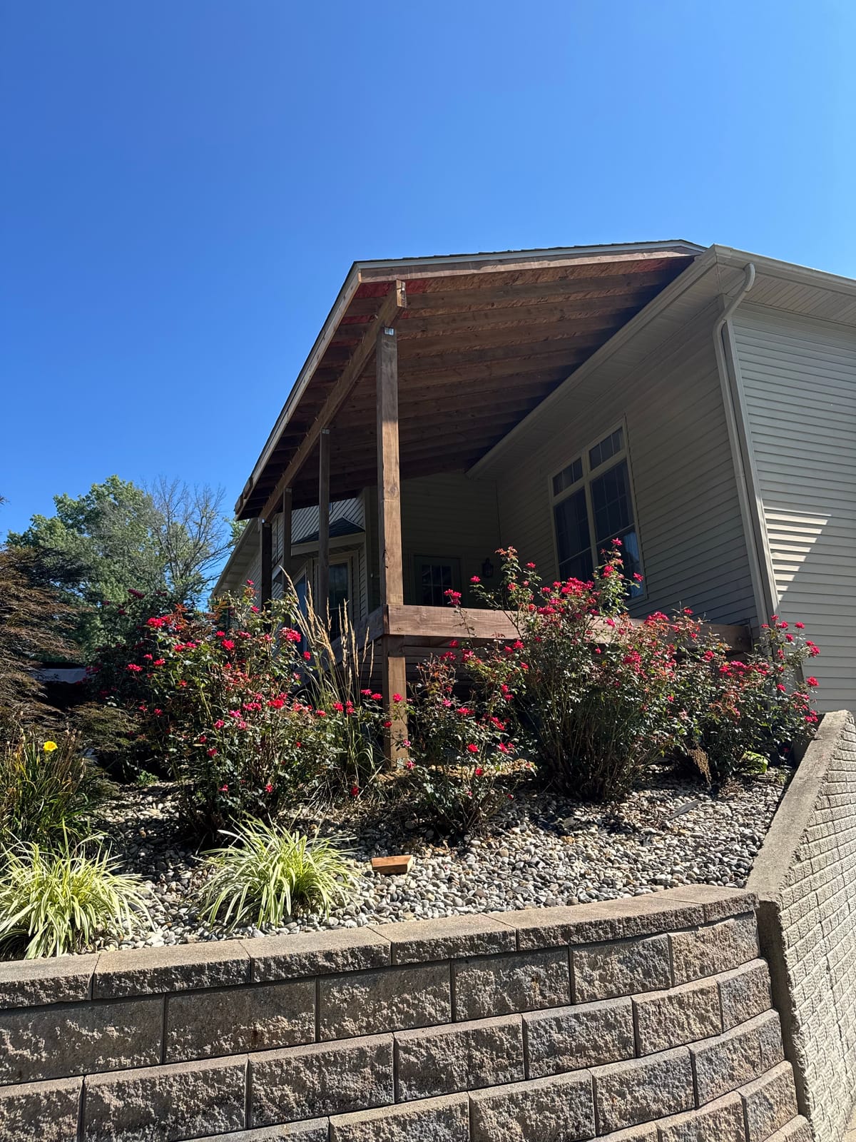 Covered porch with a stained tongue-and-groove ceiling and wood posts above red rose bushes and a block wall