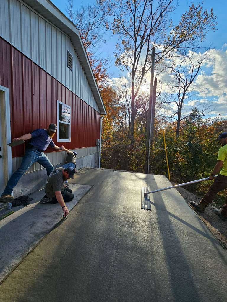 Crew finishing a fresh concrete slab with a bull float and hand trowels beside a red pole barn in fall light