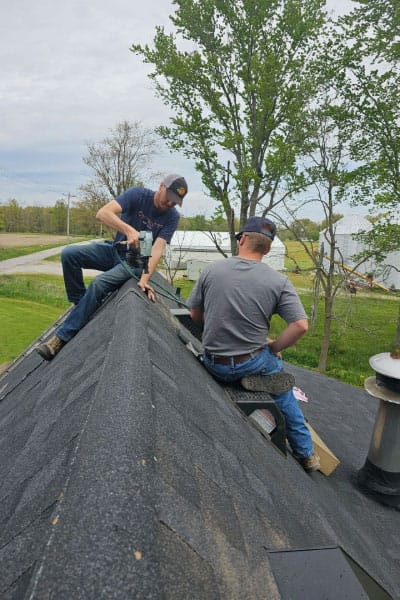 Two roofers installing ridge cap shingles with a nail gun on a dark shingle roof in the country