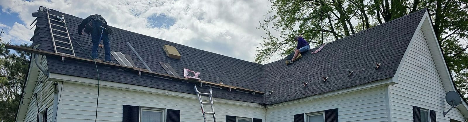 Two roofers on a dark shingle roof with roof jacks, ladders and a plank over white vinyl siding