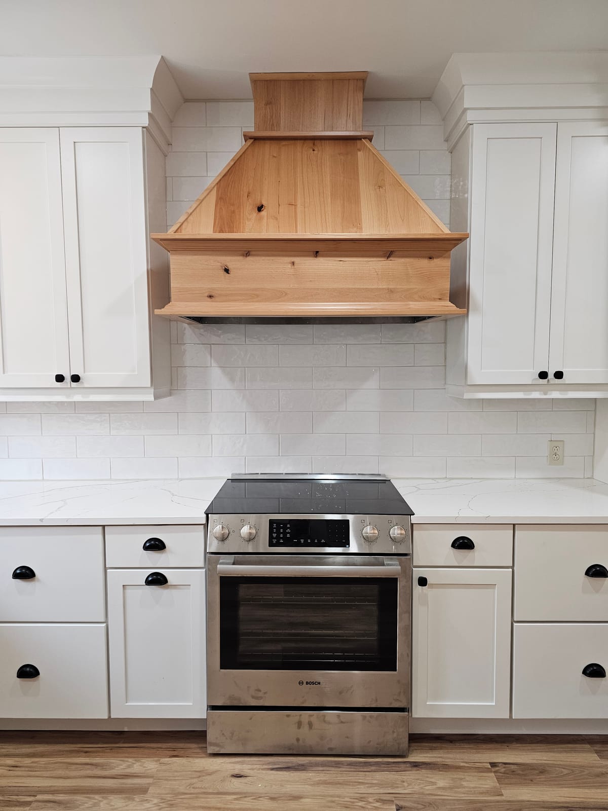 Stainless electric range under a natural knotty-wood range hood, flanked by white shaker cabinets and subway tile