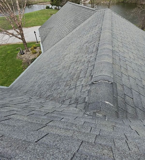Aging gray asphalt shingle roof with a split, lifting ridge cap shingle and worn granules