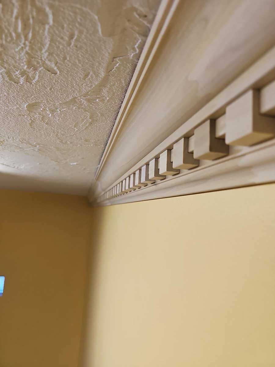 Close-up of unfinished wood crown molding with dentil blocks along a textured ceiling and yellow wall