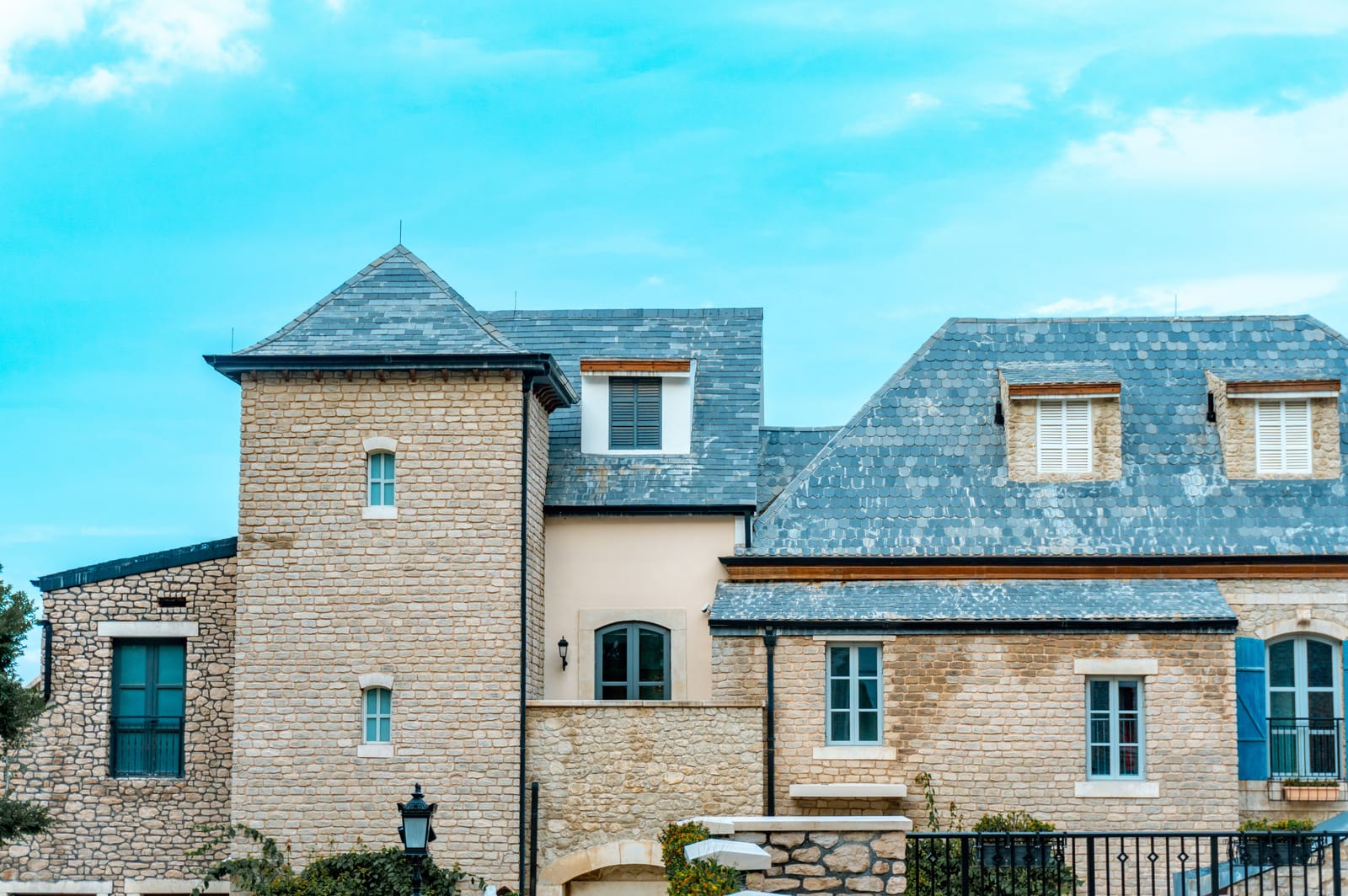 Example of stone house with a blue-gray slate-style roof, a turret and shuttered dormers under a blue sky (stock photo)