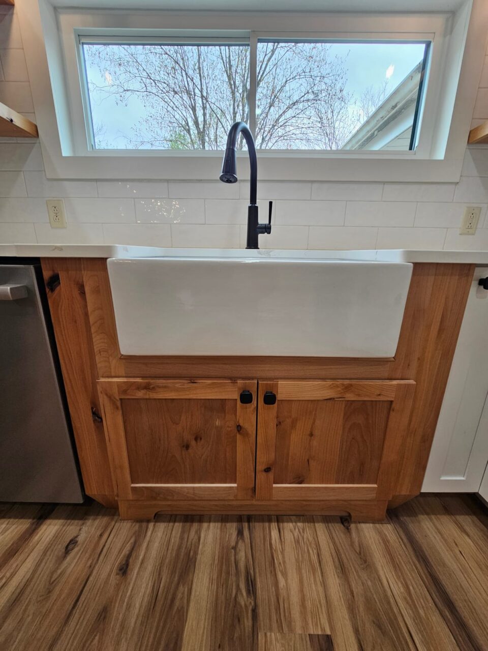 White farmhouse sink set in a knotty-wood base cabinet with black knobs, matte black faucet and a window above