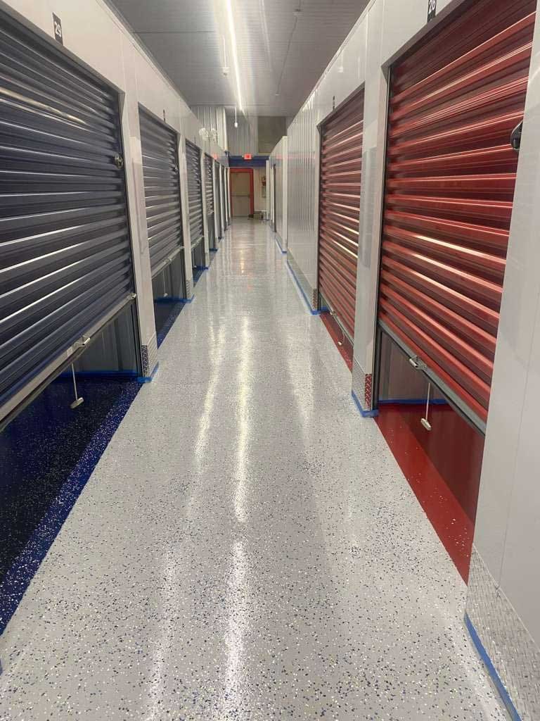 Self-storage hallway with glossy gray flake epoxy floor, roll-up doors in navy and red, and colored floor borders