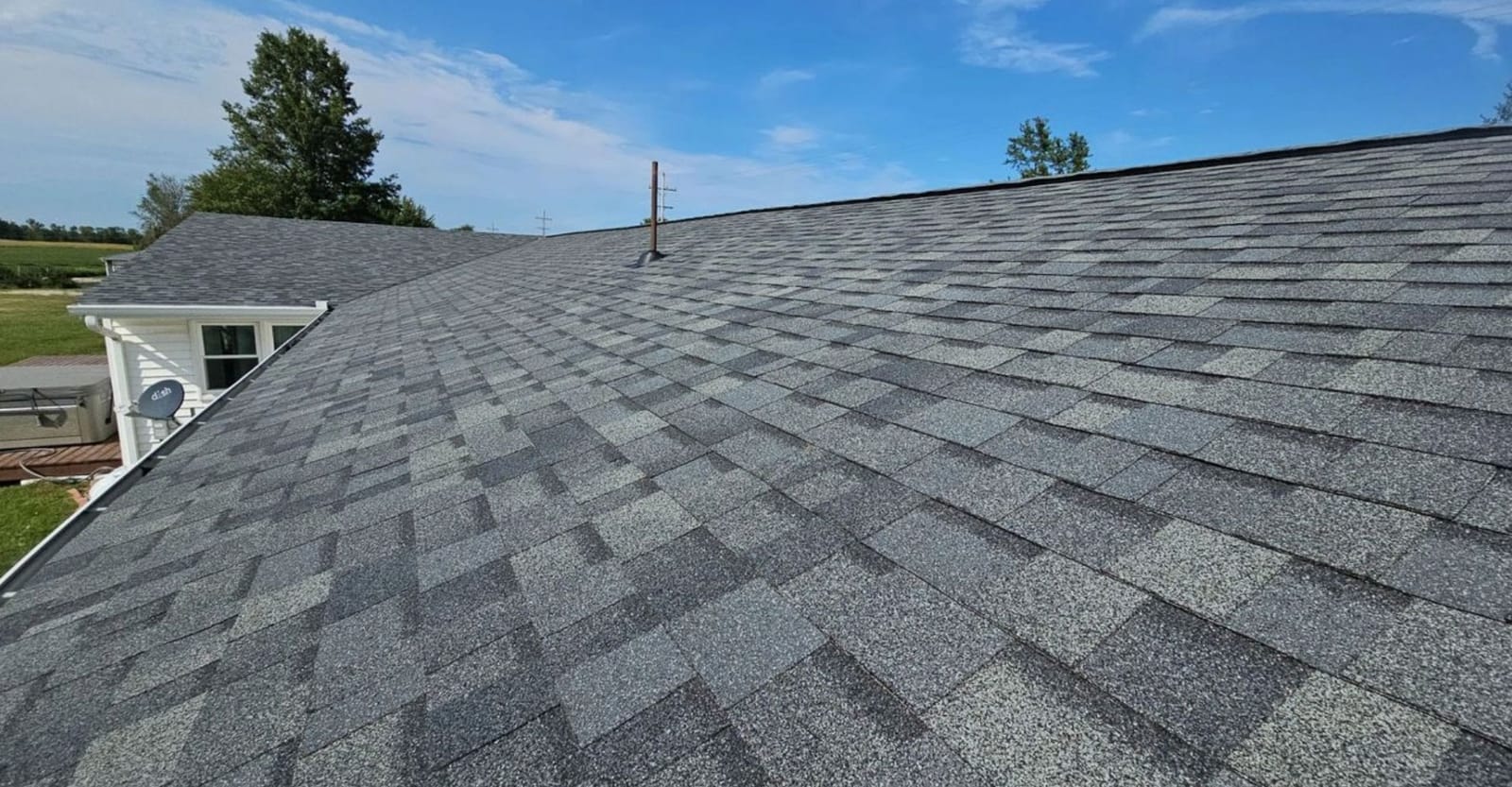 Wide view of a new gray architectural shingle roof with a pipe boot, white house wing and fields beyond