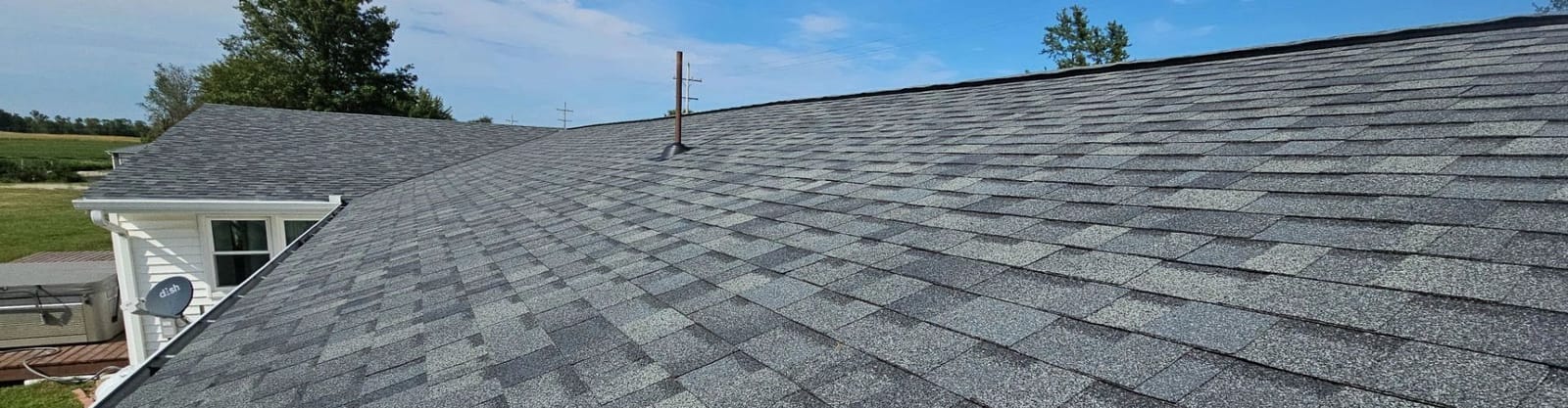 Gray architectural asphalt shingle roof with a pipe boot, white house wing and farm fields beyond