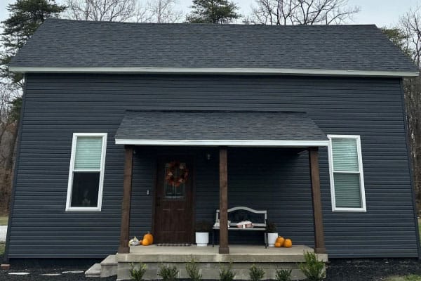 Small house with navy vinyl siding, dark shingle roof, white windows and a cedar-post covered porch