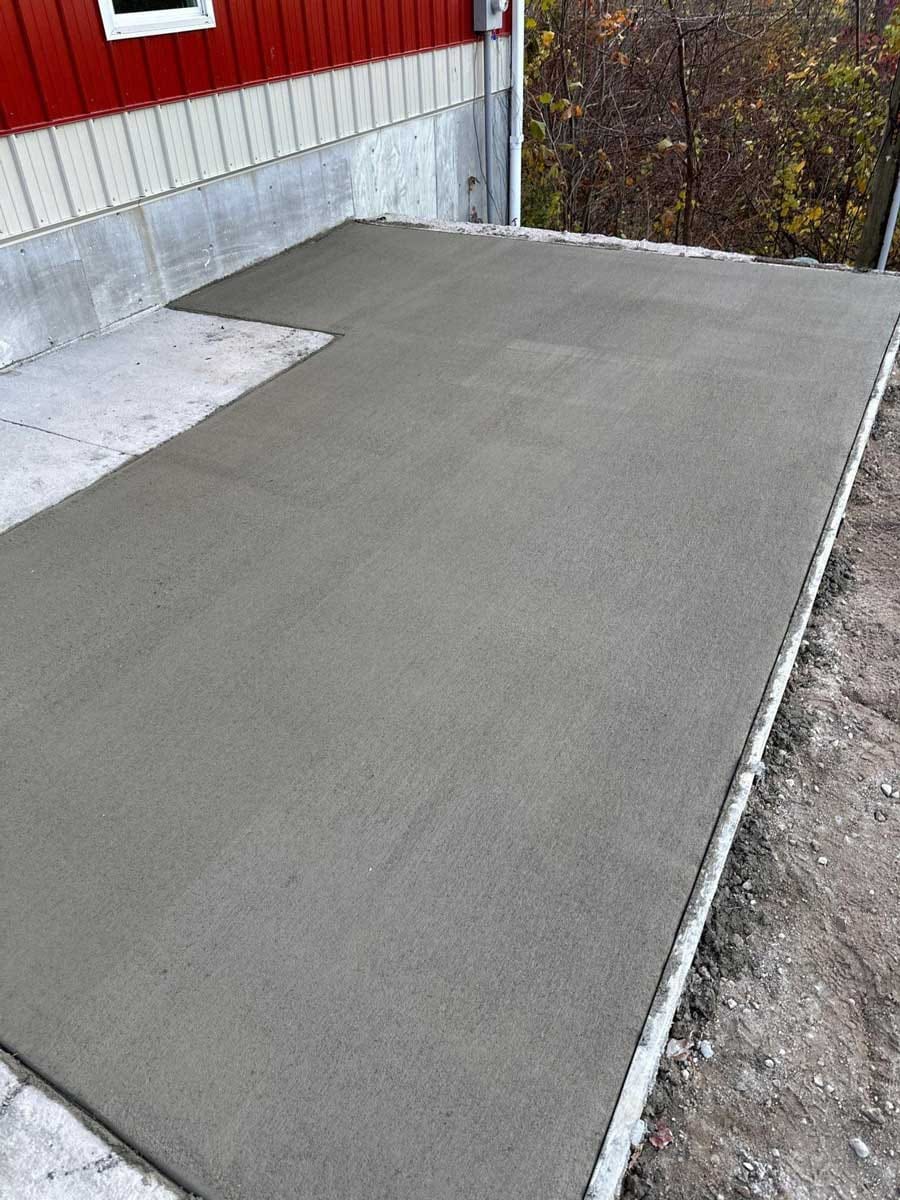 Fresh broom-finished concrete slab extending an existing pad beside a red and white metal pole building