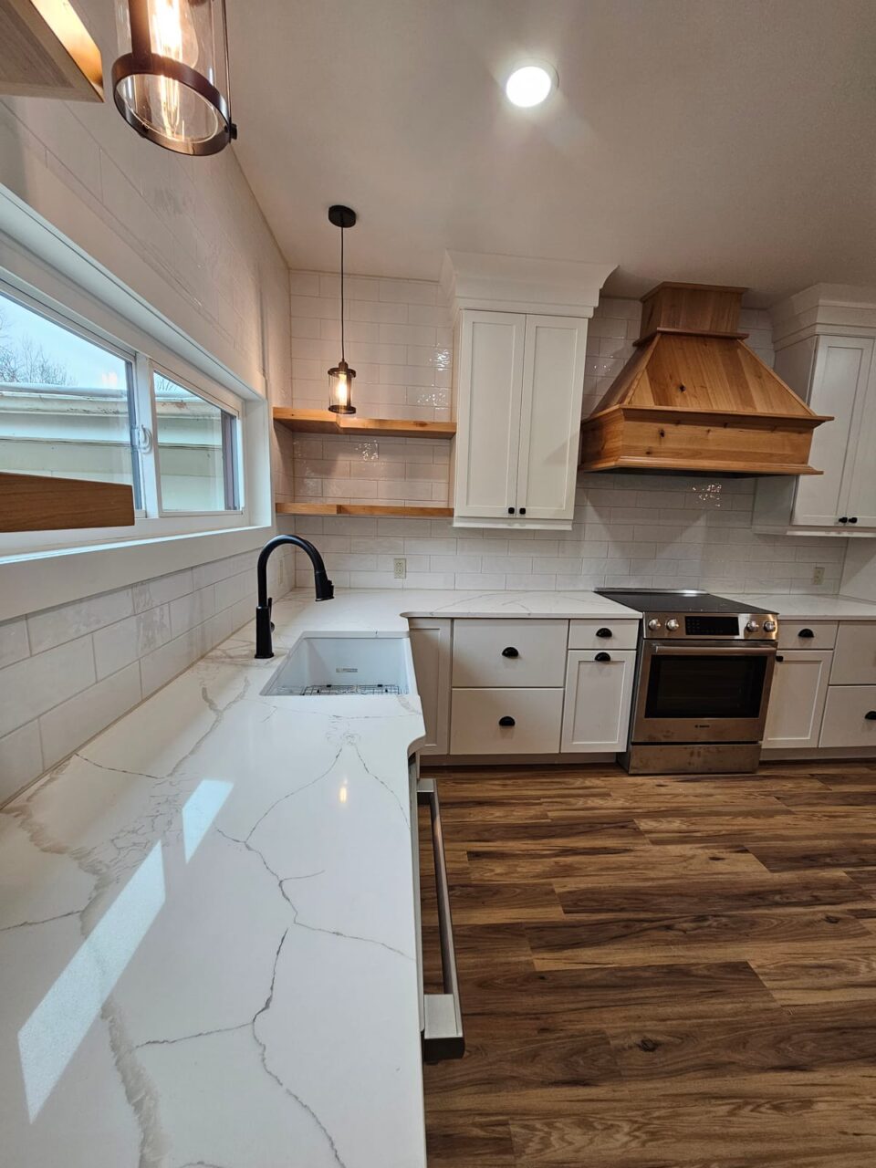 White quartz countertop with farmhouse sink and matte black faucet, white shaker cabinets and wood range hood beyond