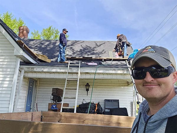 Selfie of a smiling man in sunglasses with crew tearing off an old roof on a white house behind him