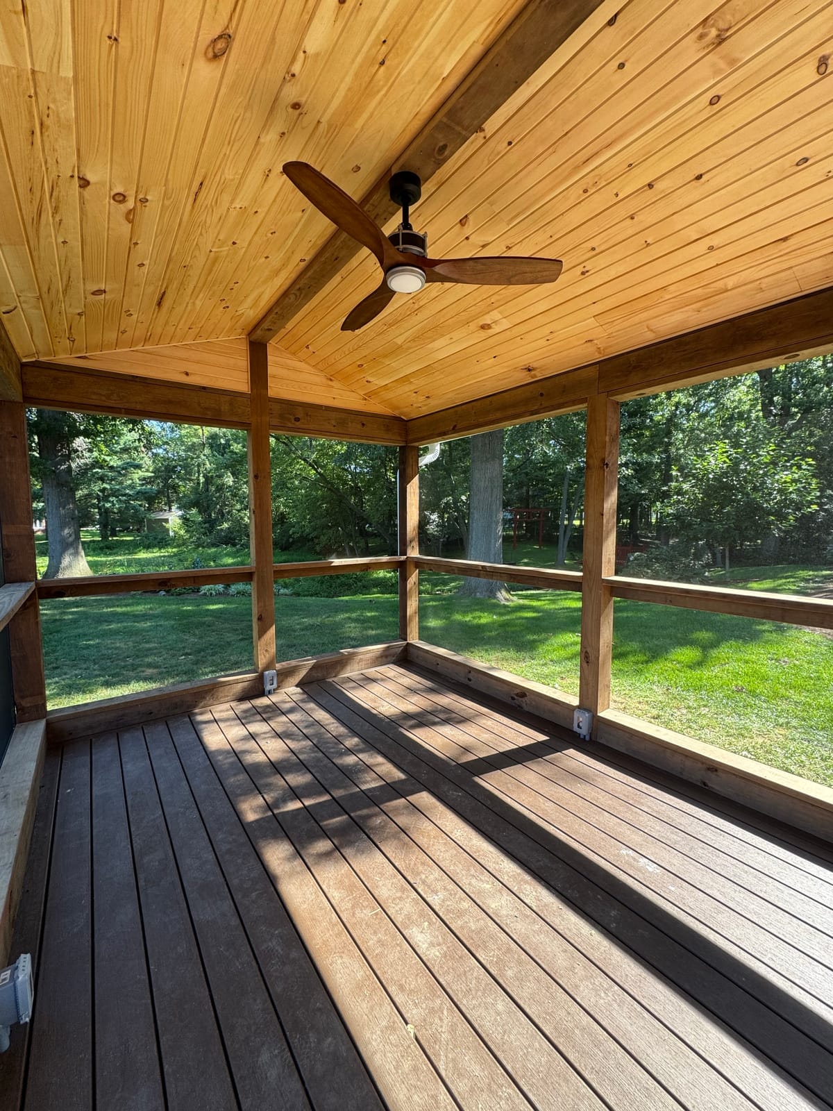 Inside a screened porch: knotty pine vaulted ceiling, wood-blade fan and brown composite floor facing a yard
