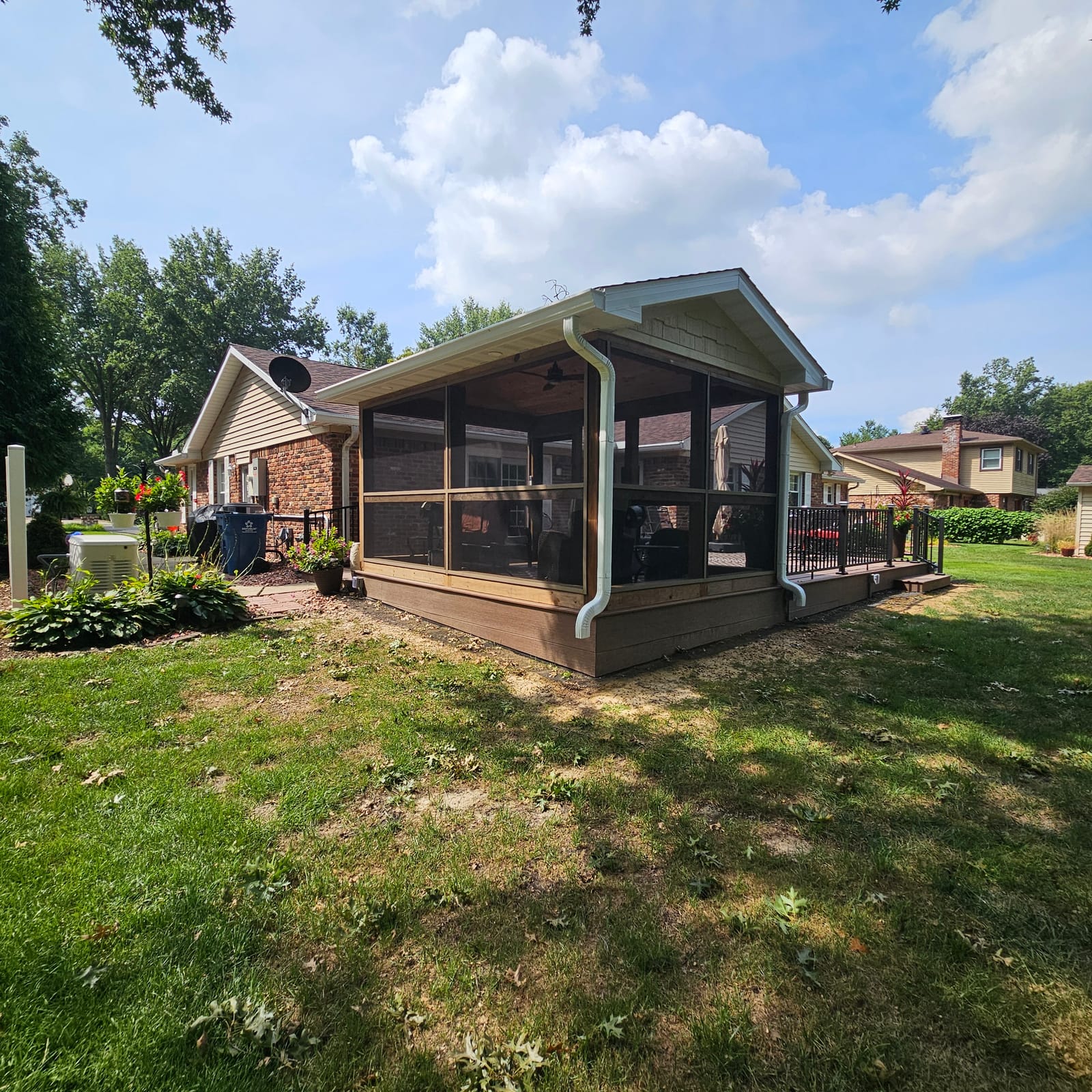 Gable-roof screened sunroom on a brown composite deck with black railing beside a brick ranch, sunny sky