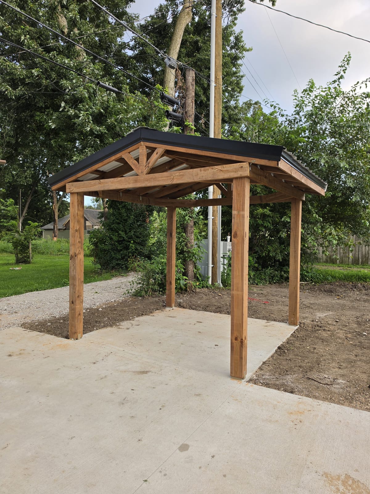 Four-post timber pavilion with a black metal roof on a new concrete slab beside a gravel drive