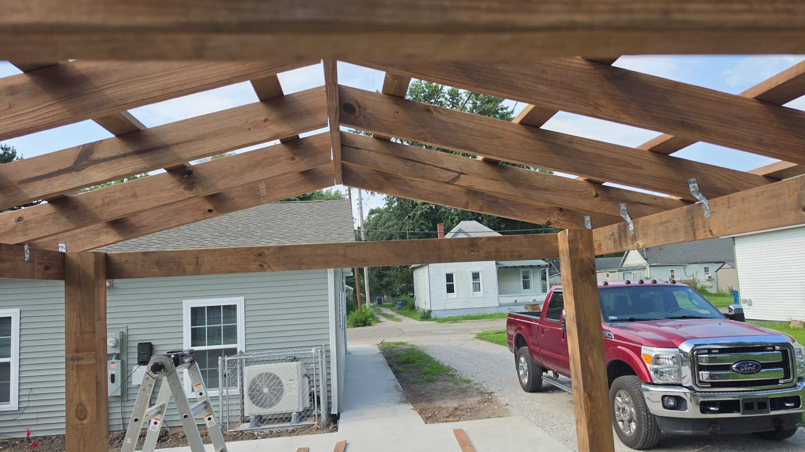 Underside of new wood pavilion rafters and beams, with a red Ford pickup and neighboring houses beyond