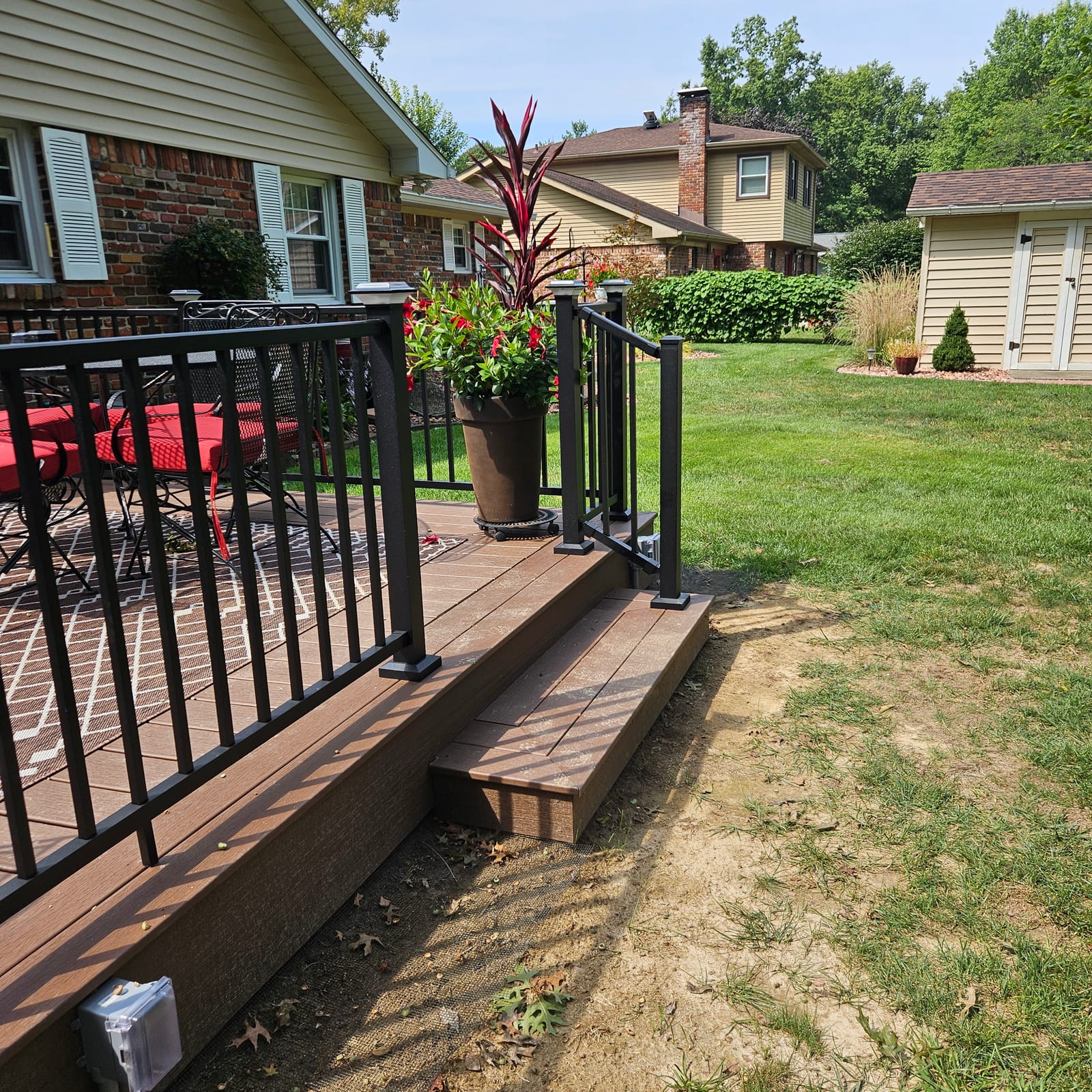Corner of a brown composite deck with black aluminum railing, box step and potted flowers on a sunny day