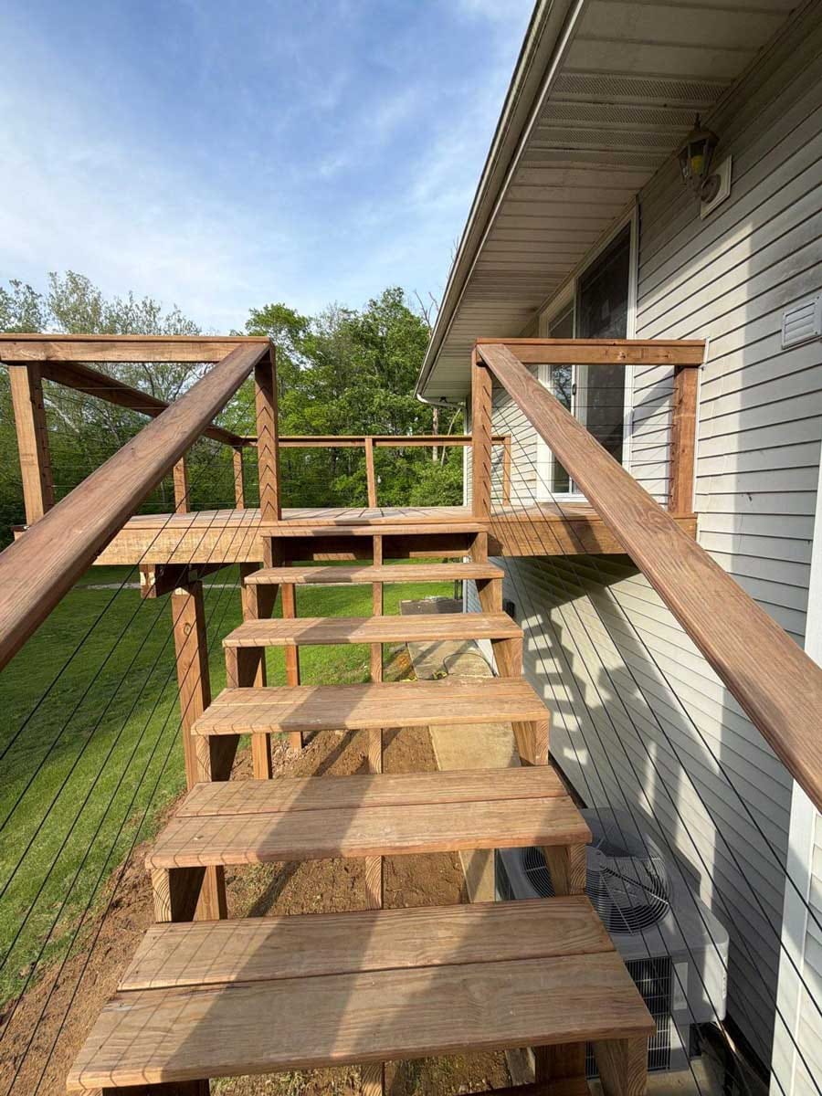 Stained wood deck stairs with black cable railing leading up to an elevated deck beside a gray-sided house