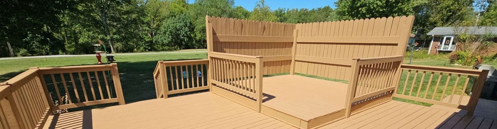 Tan solid-stained wood deck with picket railings, a raised platform and a tall privacy fence screen