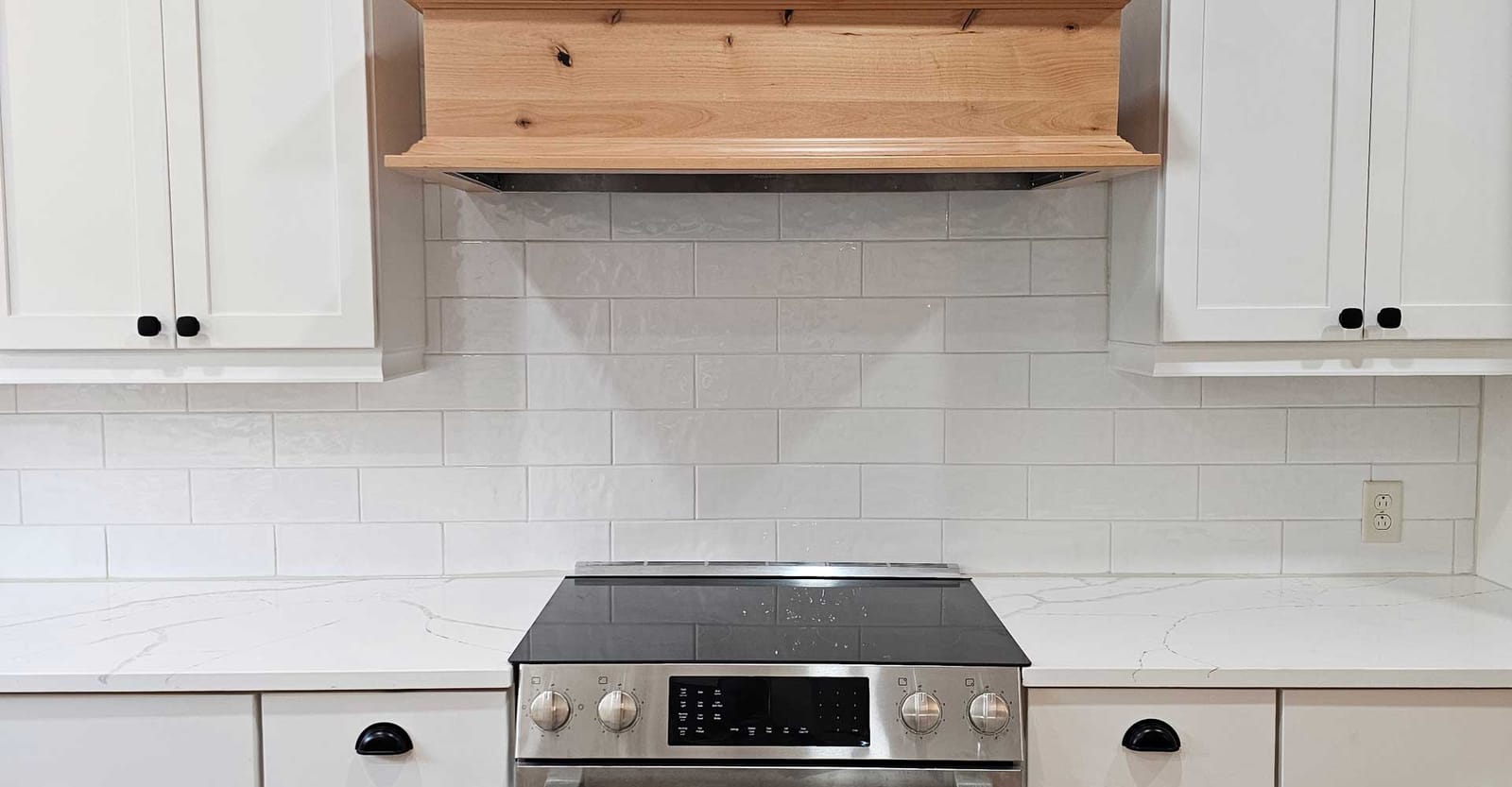Stainless electric range below a knotty-wood range hood, white subway tile, quartz counters and white shaker cabinets